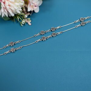 Flat lay of two 925 sterling silver anklets with detailed oxidized rose motifs and bell clusters (ghungroos) on a smooth snake chain, displayed against a solid teal surface with pink faux flowers.