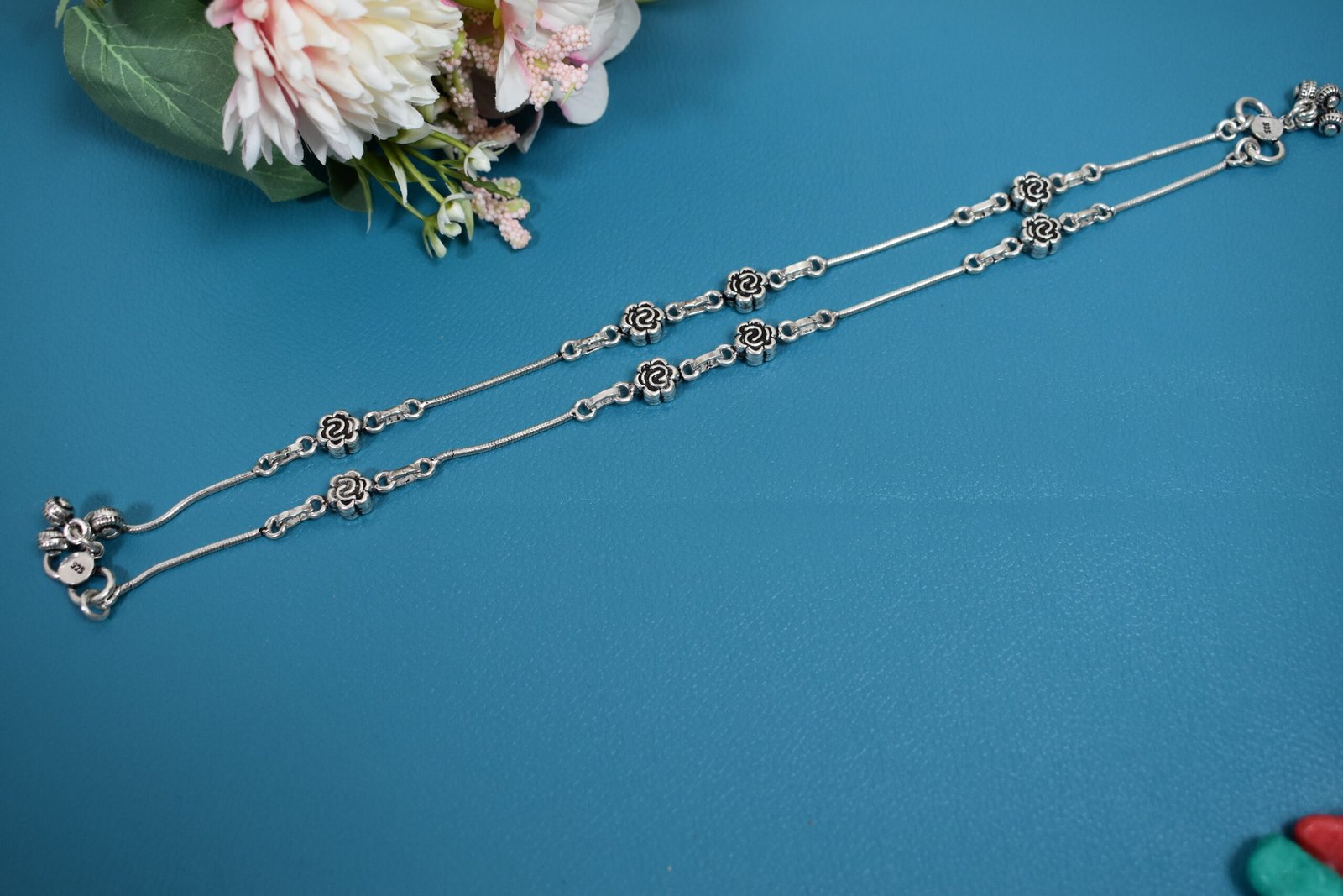 Sterling-Silver-Rose-Motif-Pair-Anklets-Teal-Background-Sukhambika-925 Flat lay of two 925 sterling silver anklets with detailed oxidized rose motifs and bell clusters (ghungroos) on a smooth snake chain, displayed against a solid teal surface with pink faux flowers.