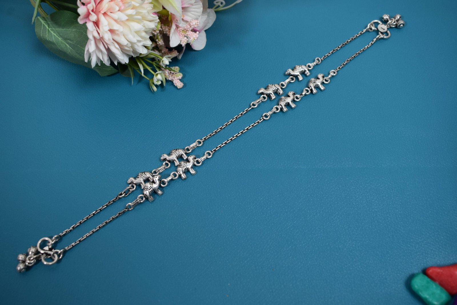 sukhambika-925-sterling-silver-elephant-charm-anklets Pair of Sukhambika 925 sterling silver anklets with multiple detailed elephant charms linked on a fine chain, resting on a textured teal background with floral and decorative accents