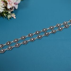 Pair of 925 sterling silver anklets featuring oxidized floral links set with clear central stones, arranged parallel on a solid teal blue background with floral accents in the corner.