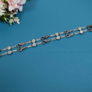 Pair of 925 sterling silver anklets featuring alternating traditional peacock motifs and textured circular links, laid parallel on a solid teal blue surface adorned with faux flowers.