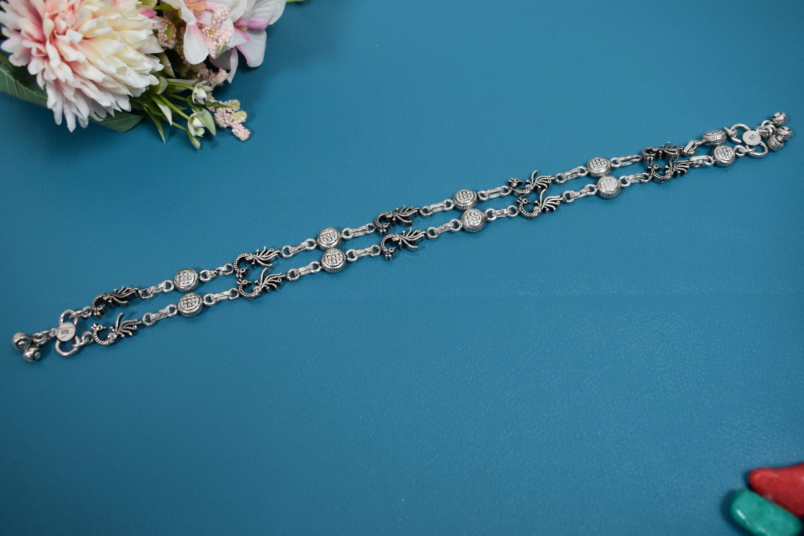925-oxidised-silver-peacock-ghungroo-anklet 2 Pair of 925 sterling silver anklets featuring alternating traditional peacock motifs and textured circular links, laid parallel on a solid teal blue surface adorned with faux flowers.