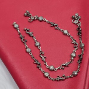 Sterling silver anklets showcasing oxidized peacock and round textured links gracefully coiled on a vibrant red textured leather surface, placed next to vintage reading glasses.