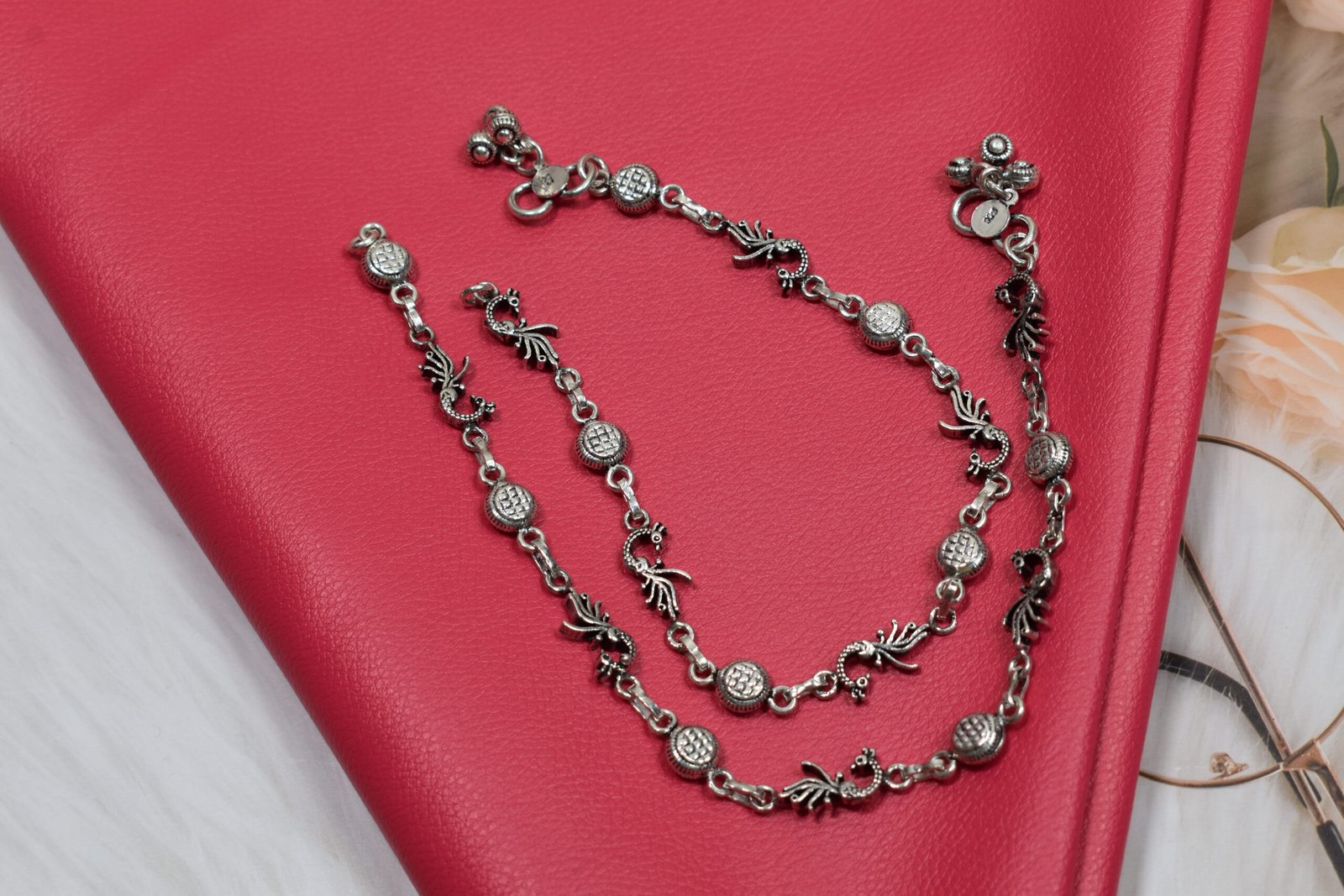 925-oxidised-silver-peacock-ghungroo-anklet3 Sterling silver anklets showcasing oxidized peacock and round textured links gracefully coiled on a vibrant red textured leather surface, placed next to vintage reading glasses.