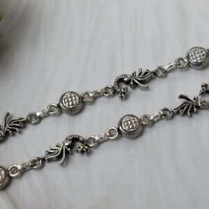 Sterling silver anklets featuring alternating oxidized peacock links and textured round discs, laid parallel on a plush white faux fur surface, highlighting the ghungroo bell clusters near the clasps.