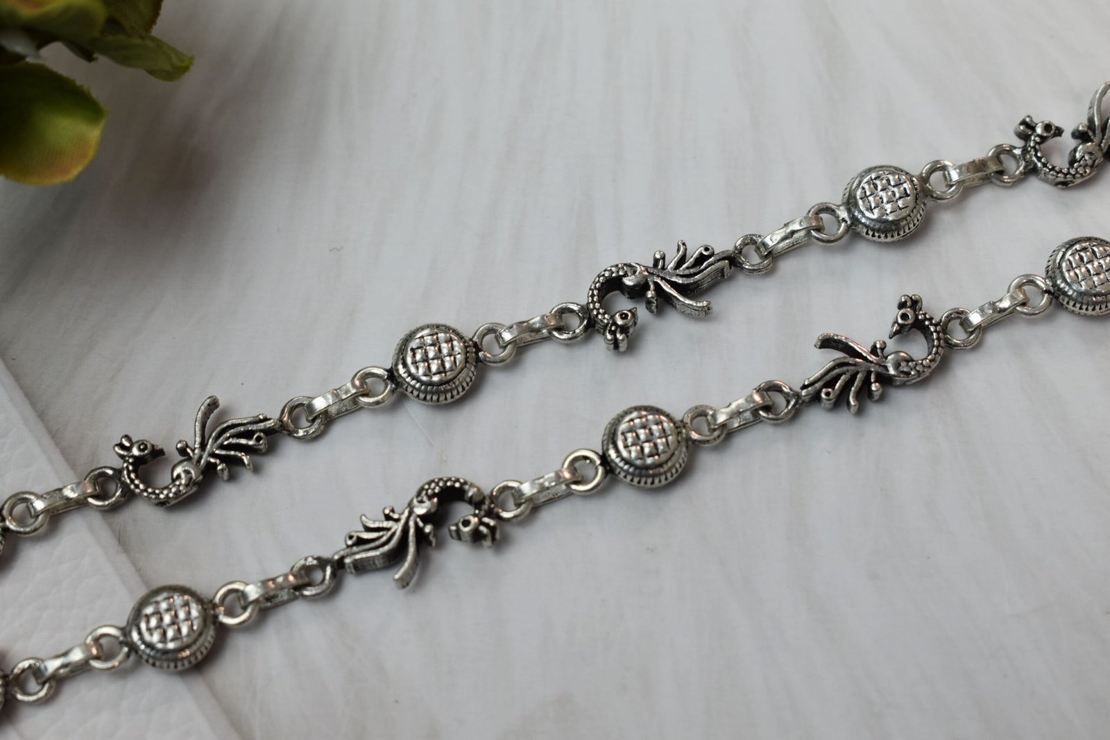 925-oxidised-silver-peacock-ghungroo-anklet5 Sterling silver anklets featuring alternating oxidized peacock links and textured round discs, laid parallel on a plush white faux fur surface, highlighting the ghungroo bell clusters near the clasps.