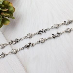 Two matching sterling silver anklets with alternating peacock and patterned disc links placed diagonally across a soft, textured white background, accented by green foliage in the corner.