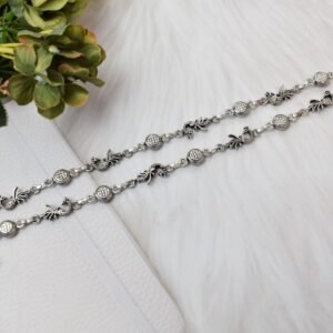 Two matching sterling silver anklets with alternating peacock and patterned disc links placed diagonally across a soft, textured white background, accented by green foliage in the corner.
