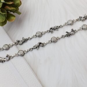 Two matching sterling silver anklets with alternating peacock and patterned disc links placed diagonally across a soft, textured white background, accented by green foliage in the corner.