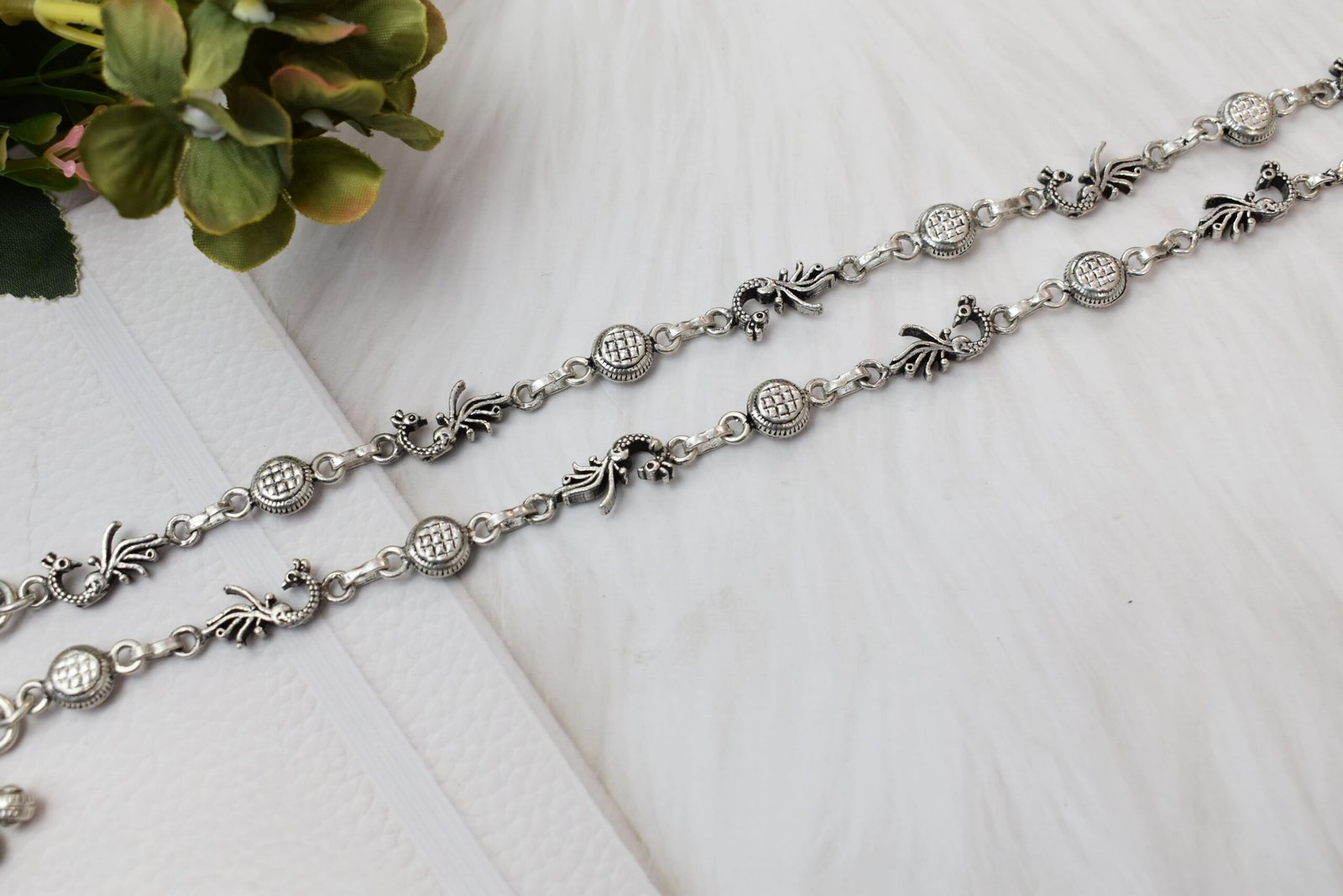 925-oxidised-silver-peacock-ghungroo-anklet9 Two matching sterling silver anklets with alternating peacock and patterned disc links placed diagonally across a soft, textured white background, accented by green foliage in the corner.