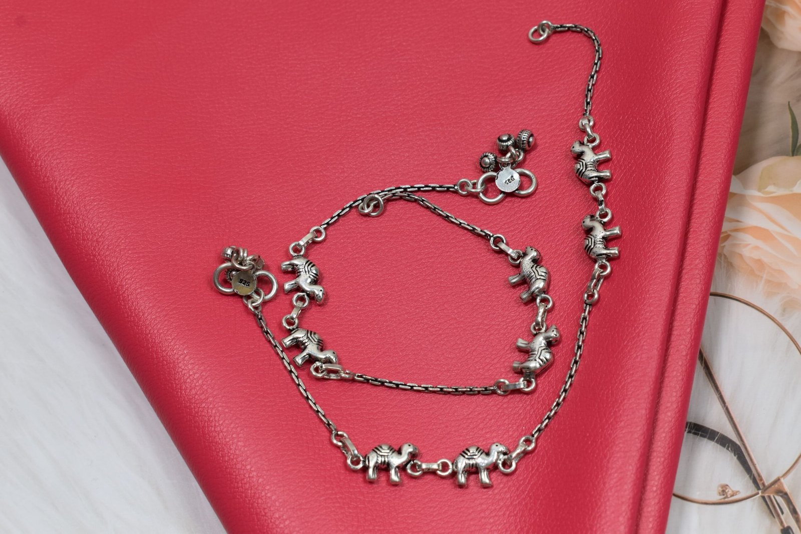 sukhambika-925-sterling-silver-elephant-charm-anklets Pair of Sukhambika 925 sterling silver anklets with multiple detailed elephant charms linked on a fine chain, resting on a textured teal background with floral and decorative accents