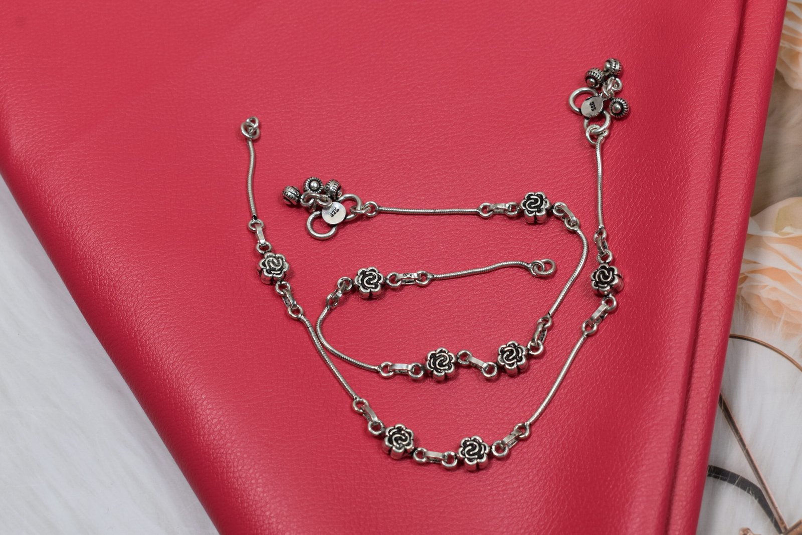 Oxidized-Rose-Silver-Anklet-with-Large-Ghungroo-Red-Background-Sukhambika-925 Close-up of a single 925 sterling silver anklet featuring detailed oxidized rose links and a cluster of larger, textured ghungroo bells on a smooth snake chain, resting on red textured leather. A clear '925' stamp is visible near the clasp