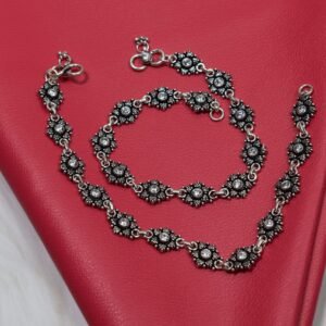 A pair of sterling silver anklets with stone-studded floral links elegantly coiled on a textured red surface, accompanied by vintage reading glasses.