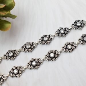 Close-up shot of two sterling silver anklets displaying detailed oxidized floral motifs with sparkling clear stones in the center, laid out diagonally on a white textured surface next to a green plant.