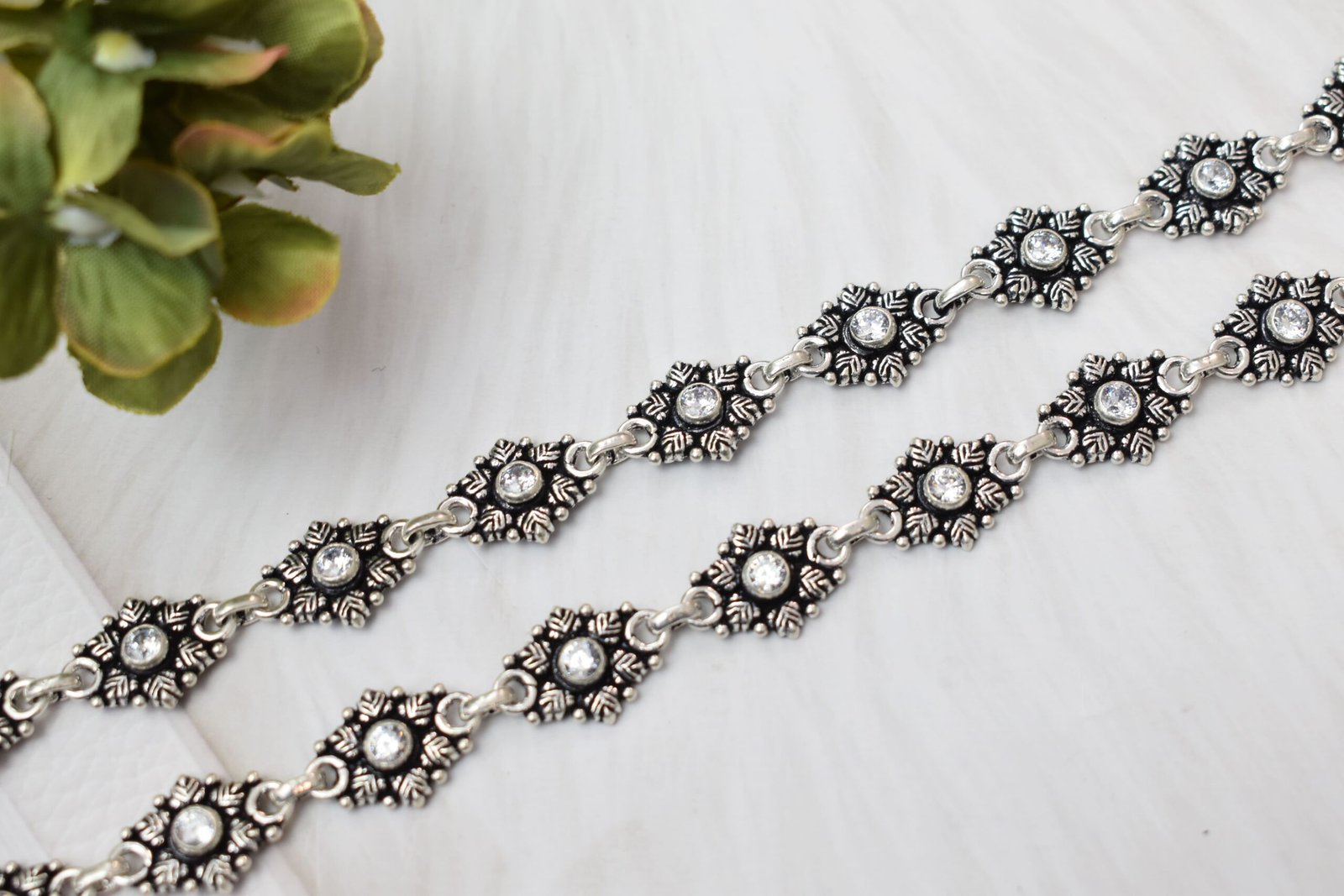 Close-up-Stone-Studded-Silver-Anklet-Links-Sukhambika-925 Close-up shot of two sterling silver anklets displaying detailed oxidized floral motifs with sparkling clear stones in the center, laid out diagonally on a white textured surface next to a green plant.
