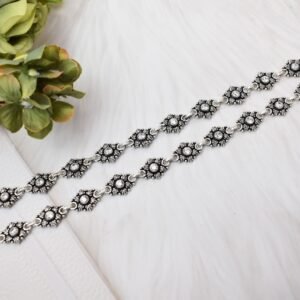 Two matching 925 sterling silver anklets with clear stones set in floral oxidized links, laid straight on a soft white surface accompanied by green and pink botanical accents.
