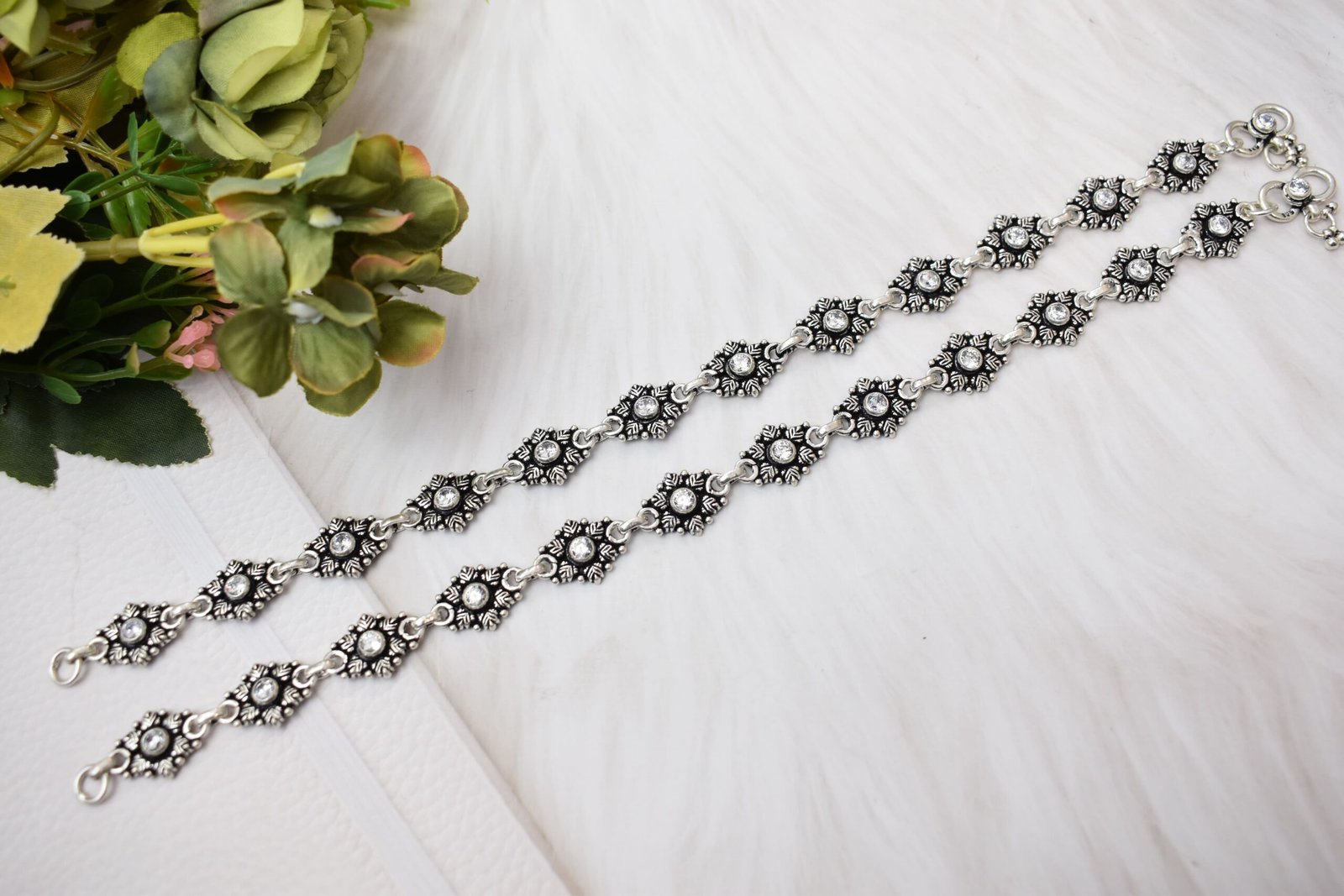 Elegant-Stone-Floral-Silver-Anklets-White-Surface-Sukhambika-925 Two matching 925 sterling silver anklets with clear stones set in floral oxidized links, laid straight on a soft white surface accompanied by green and pink botanical accents.