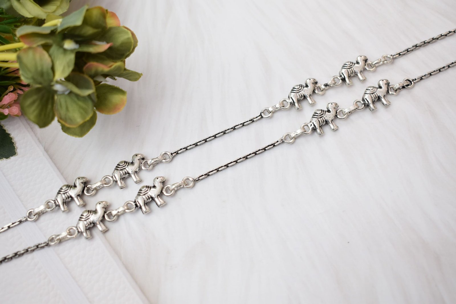 sukhambika-925-sterling-silver-elephant-charm-anklets Pair of Sukhambika 925 sterling silver anklets with multiple detailed elephant charms linked on a fine chain, resting on a textured teal background with floral and decorative accents