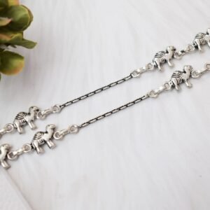 Pair of Sukhambika 925 sterling silver anklets with multiple detailed elephant charms linked on a fine chain, resting on a textured teal background with floral and decorative accents.