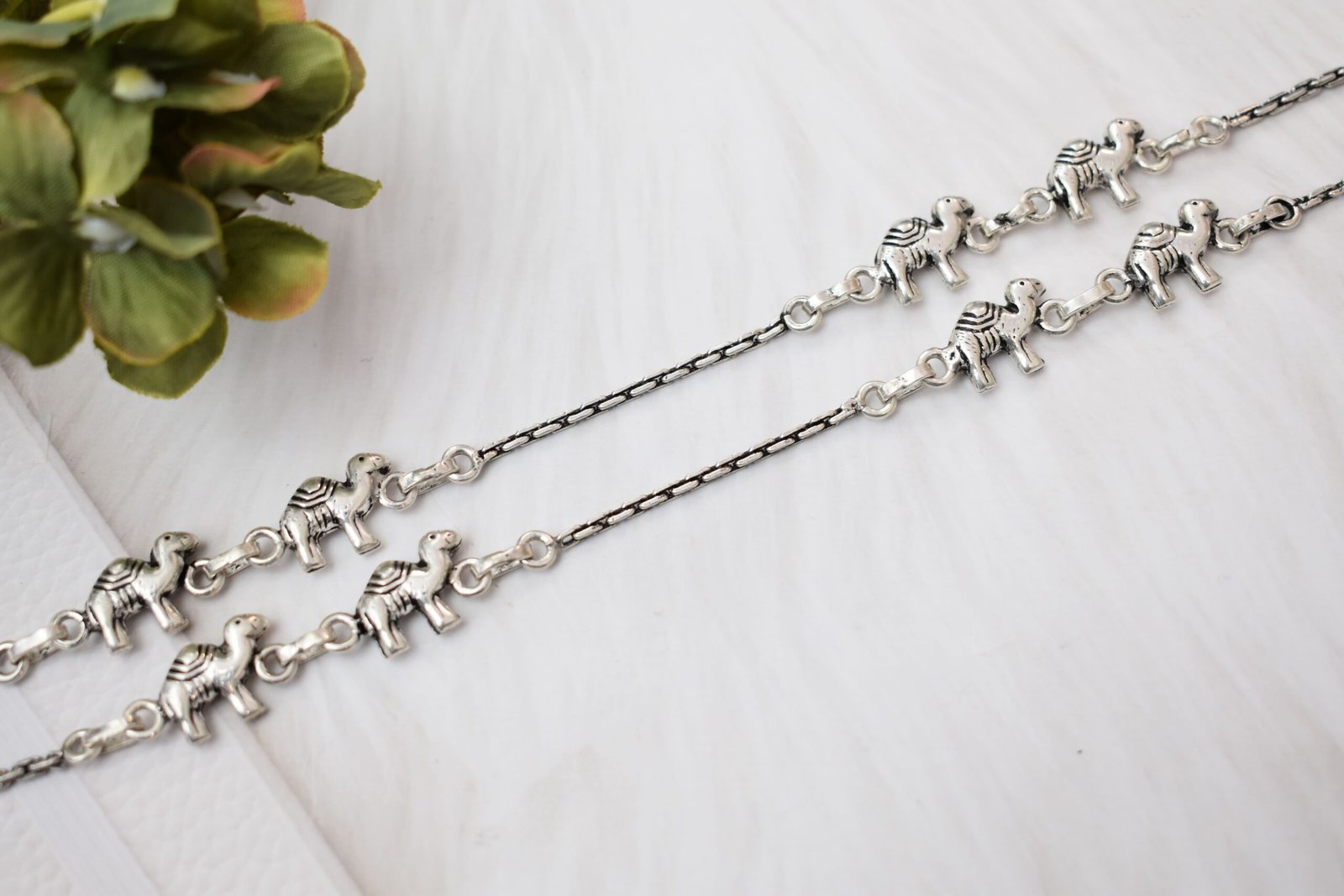 sukhambika-925-sterling-silver-elephant-charm-anklets. Pair of Sukhambika 925 sterling silver anklets with multiple detailed elephant charms linked on a fine chain, resting on a textured teal background with floral and decorative accents.