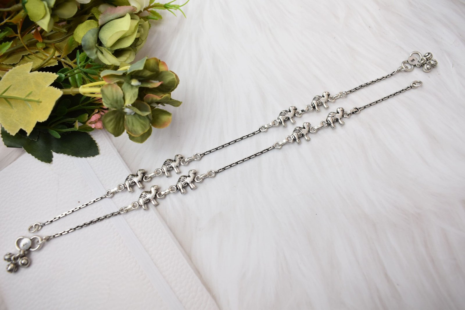 sukhambika-925-sterling-silver-elephant-charm-anklets Pair of Sukhambika 925 sterling silver anklets with multiple detailed elephant charms linked on a fine chain, resting on a textured teal background with floral and decorative accents