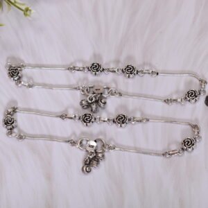 Flat lay of two matching 925 sterling silver anklets, laid parallel, featuring spaced rose motifs and large, textured central ghungroo bell clusters on smooth chains, resting on white plush faux fur next to vintage reading glasses and leaves.