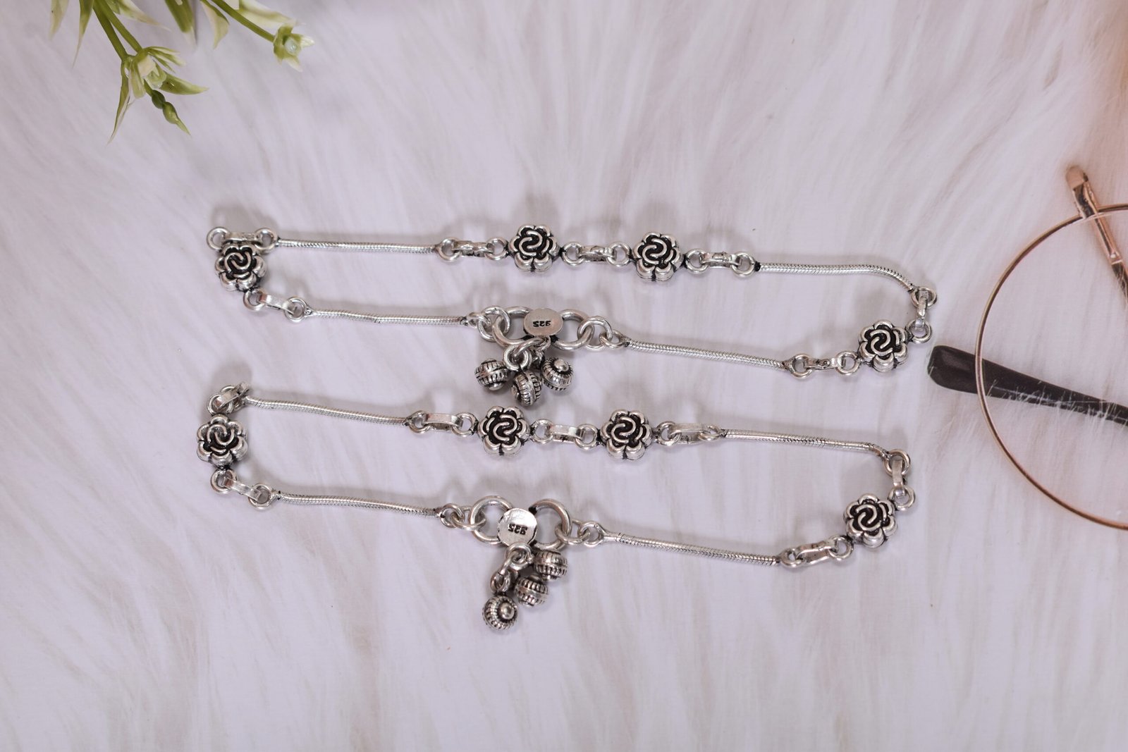 Vintage-Style-Silver-Anklets-Pair-Ghungroo-Central-Cluster-White-Fur-Sukhambika-925 Flat lay of two matching 925 sterling silver anklets, laid parallel, featuring spaced rose motifs and large, textured central ghungroo bell clusters on smooth chains, resting on white plush faux fur next to vintage reading glasses and leaves.