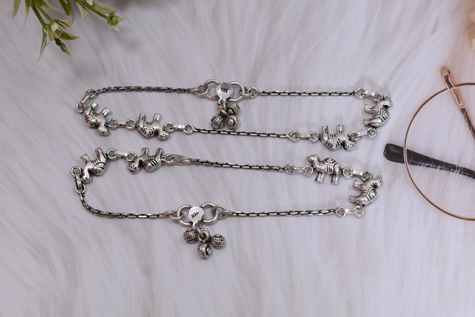 sukhambika-925-sterling-silver-elephant-charm-anklets Pair of Sukhambika 925 sterling silver anklets with multiple detailed elephant charms linked on a fine chain, resting on a textured teal background with floral and decorative accents