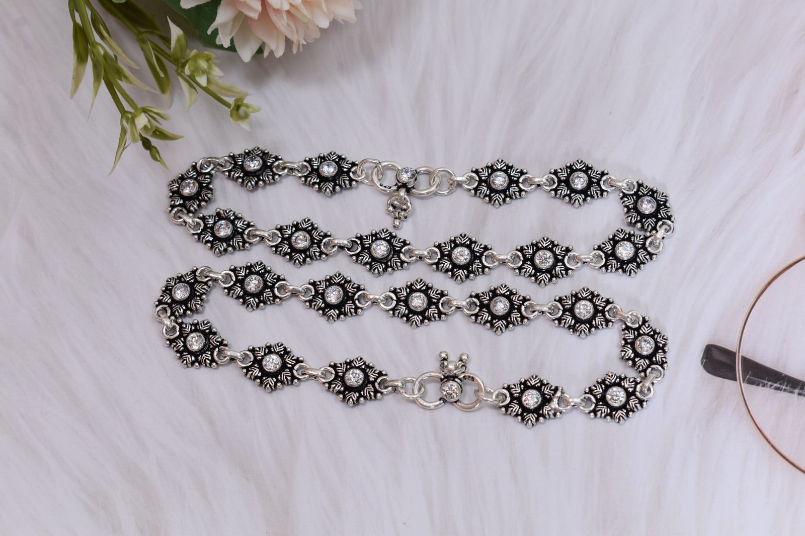 Stone-Studded-Floral-Silver-Anklets-Coiled-Sukhambika-925 Pair of 925 sterling silver anklets coiled gracefully on a fluffy white faux fur surface, highlighting the clear center stones in oxidized floral settings and a visible 925 stamp on the S-hook clasp.