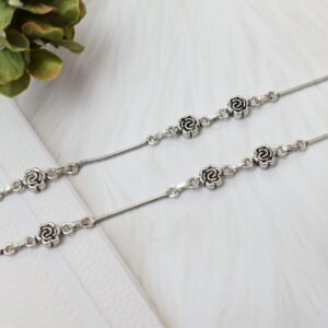 A macro close-up photograph focusing on the precise joinery between a single oxidized silver rose flower link and the smooth silver snake chain of an anklet, set against a soft, textured white background.