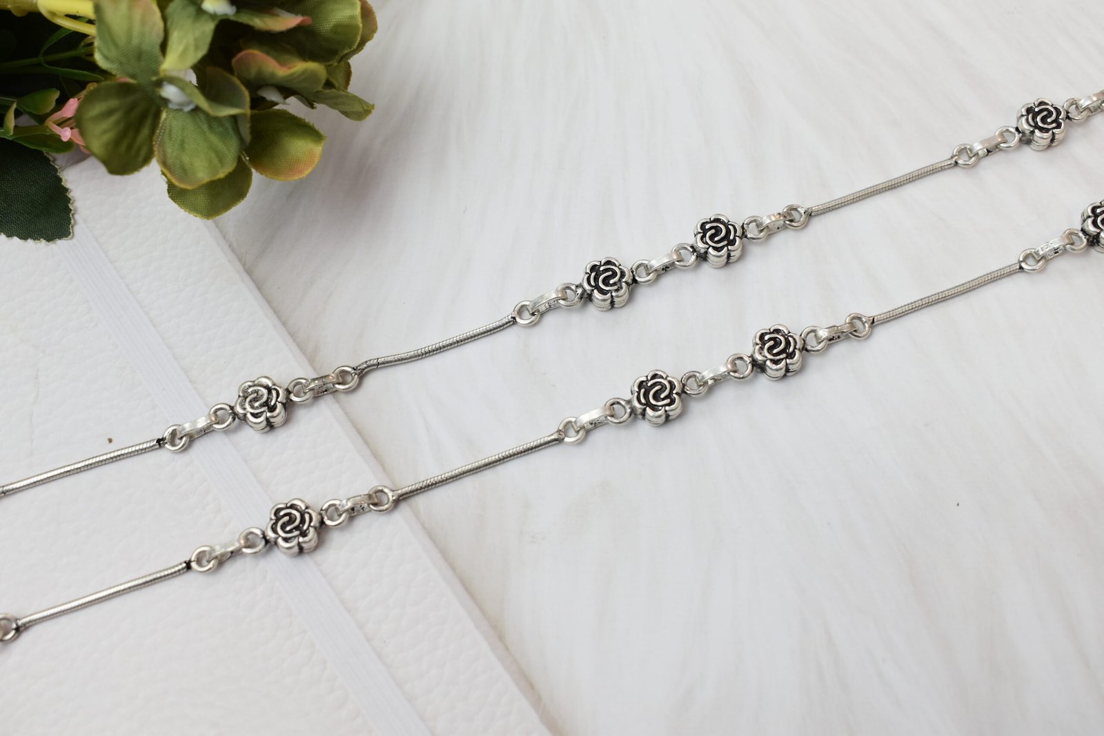 Close-up-Craftsmanship-Rose-Link-Silver-Anklet-White-Surface-Sukhambika-925 A macro close-up photograph focusing on the precise joinery between a single oxidized silver rose flower link and the smooth silver snake chain of an anklet, set against a soft, textured white background.