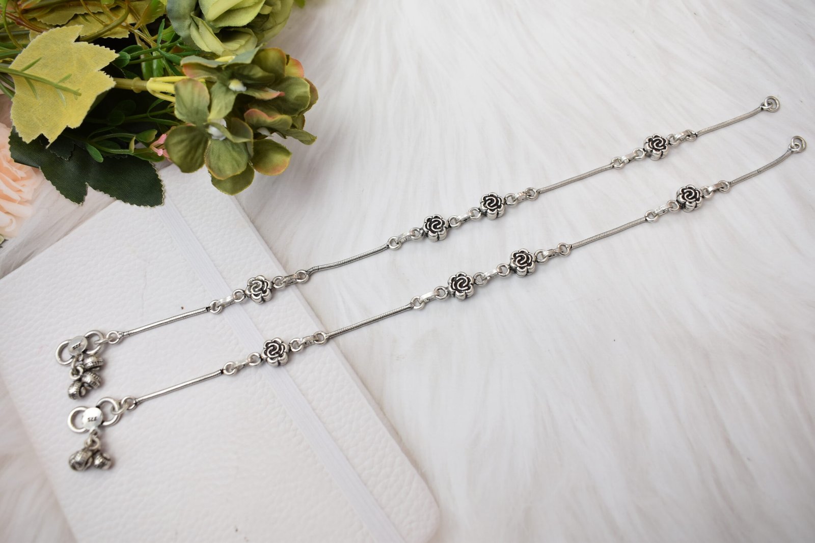 Complete-Pair-Rose-Anklets-Teal-Teal-Blue-Surface-Sukhambika-925 Top-down view of two parallel 925 sterling silver anklets, featuring spaced rose motifs, small spherical ghungroo bells near the clasps, and prominent '925' stamps, laid out in parallel on a textured teal blue background with faux flowers.