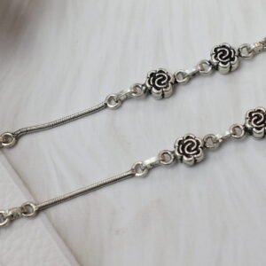 Close-up of a 925 sterling silver anklet featuring intricate oxidized rose flower links spaced along a smooth silver snake chain, resting on a textured white surface.