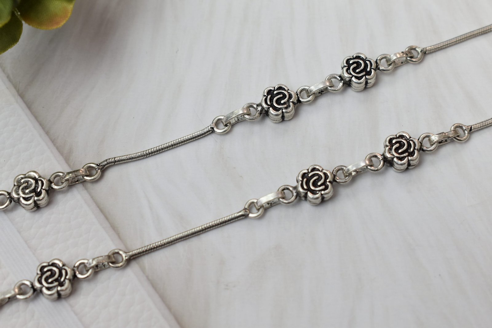 Sterling-Silver-Rose-Motif-Anklet-Sukhambika-925 Close-up of a 925 sterling silver anklet featuring intricate oxidized rose flower links spaced along a smooth silver snake chain, resting on a textured white surface.