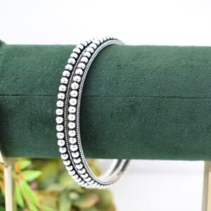 A pair of 925 sterling silver kadas overlapping on a dark green velvet jewelry stand, featuring a continuous silver ball fusion pattern bordered by a delicate twisted rope design.