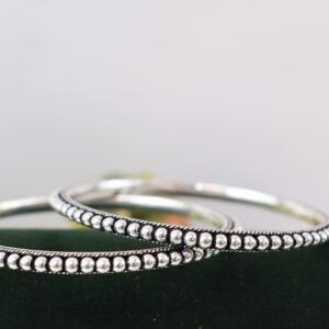 A pair of 925 sterling silver kadas overlapping on a dark green velvet jewelry stand, featuring a continuous silver ball fusion pattern bordered by a delicate twisted rope design.