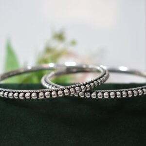A pair of 925 sterling silver kadas overlapping on a dark green velvet jewelry stand, featuring a continuous silver ball fusion pattern bordered by a delicate twisted rope design.