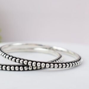 A pair of 925 sterling silver kadas overlapping on a dark green velvet jewelry stand, featuring a continuous silver ball fusion pattern bordered by a delicate twisted rope design.