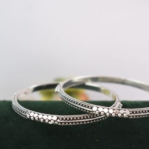 A pair of 925 sterling silver kadas overlapping on a dark green velvet jewelry stand, showcasing an intricate barik dana (fine bead) pattern along the oxidized bands.