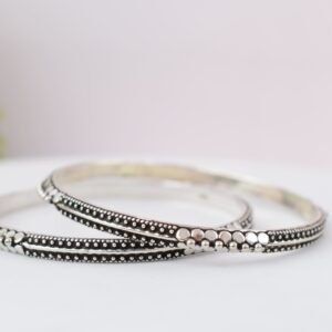 A pair of 925 sterling silver kadas overlapping on a dark green velvet jewelry stand, showcasing an intricate barik dana (fine bead) pattern along the oxidized bands.