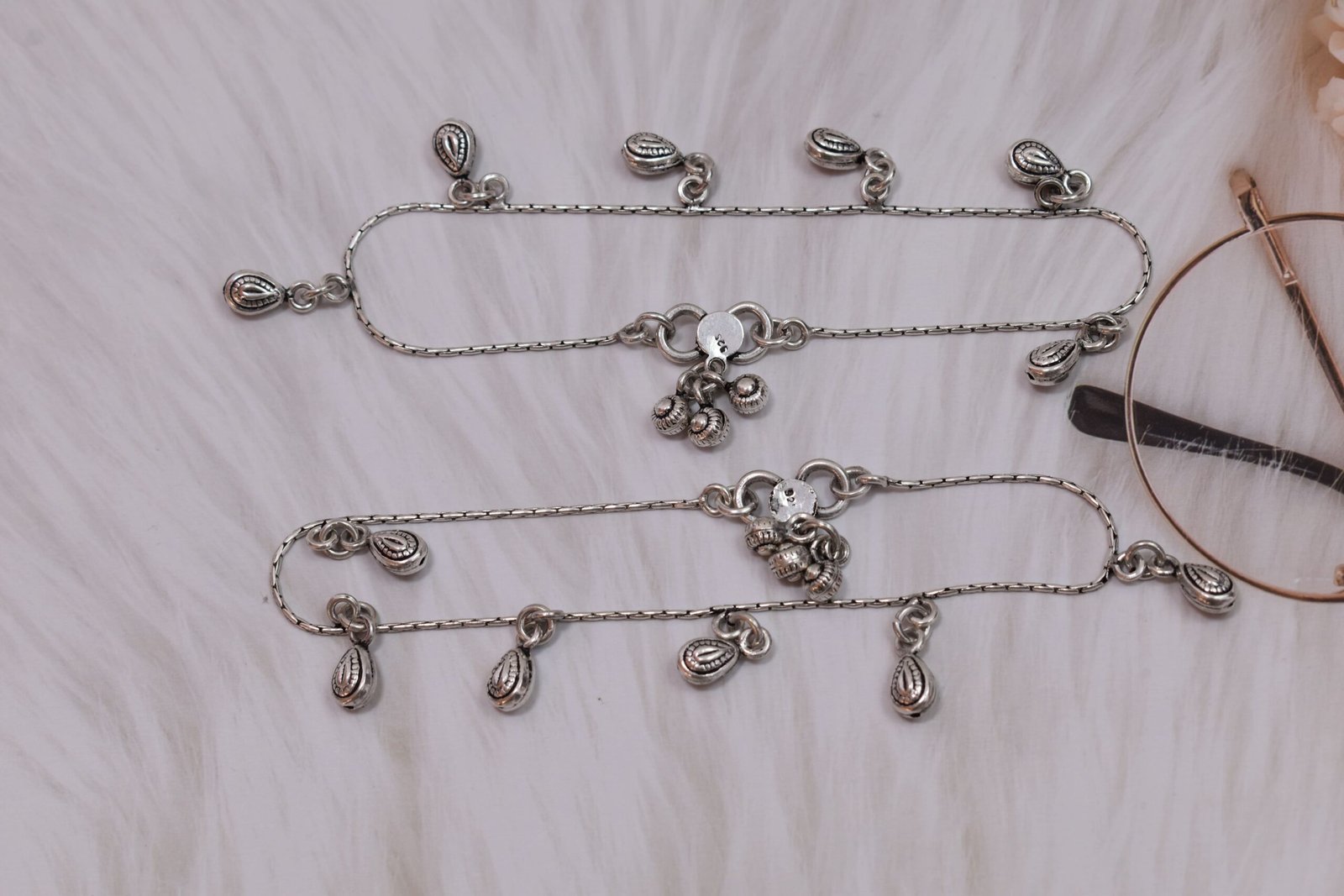classic-chained-beads-drop-silver-anklets3 A full-length view of a pair of classic chained beads drop silver anklets stretched out on a white surface next to floral accents, showing the complete chains, drop charms, and ghungroo clasps.