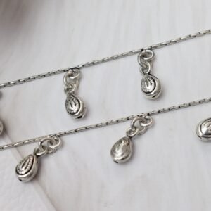 Macro close-up photograph of classic chained beads drop silver anklets, highlighting the detailed, textured engravings on the dangling teardrop charms against a soft white surface.