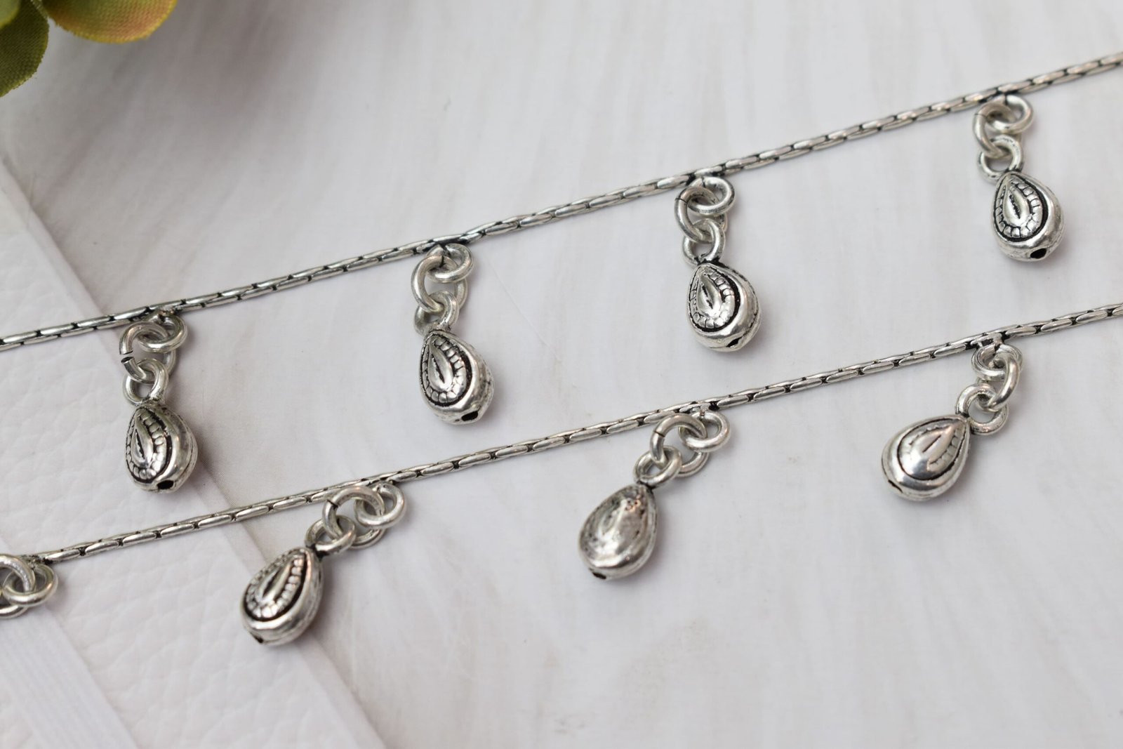 Close-Up-Textured-Bead-Drop-Silver-Anklet-Sukhambika-925 Macro close-up photograph of classic chained beads drop silver anklets, highlighting the detailed, textured engravings on the dangling teardrop charms against a soft white surface.