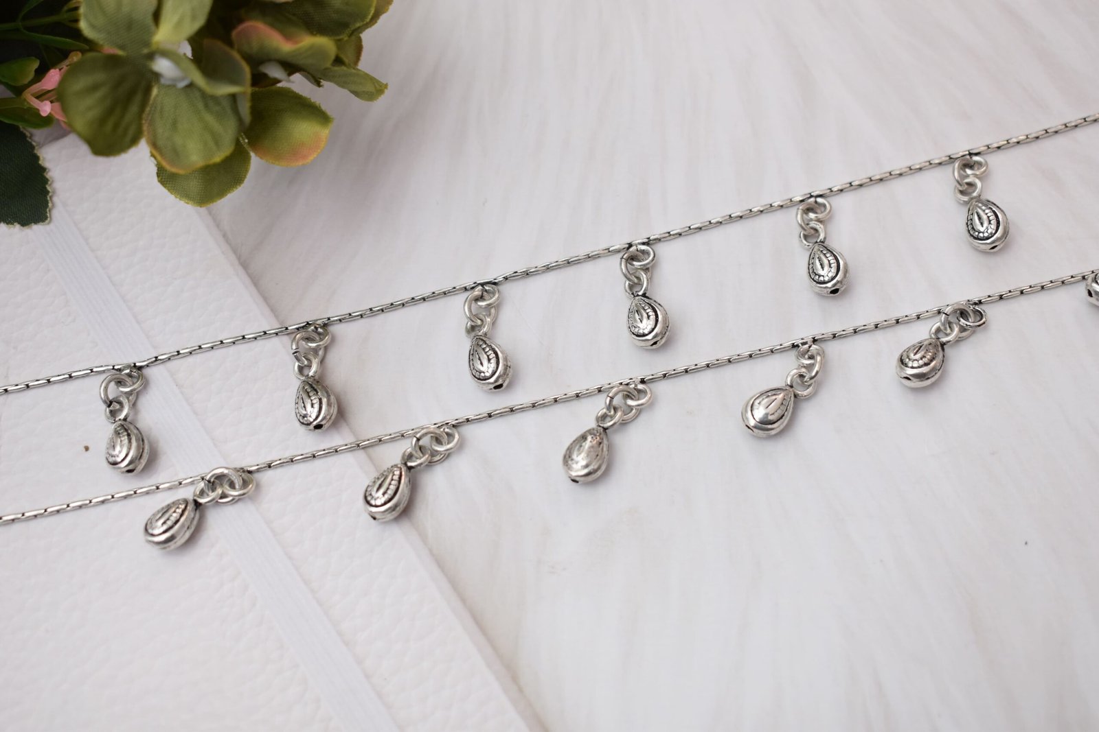 classic-chained-beads-drop-silver-anklets6 Close-up diagonal view of two parallel silver chains featuring multiple hanging textured bead drops, resting across a soft white textured backdrop.