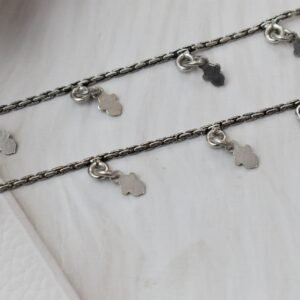 a pair of classic chained leaf shape drop 925 sterling silver anklets stretched across a white textured surface and white faux fur, featuring flat leaf charms and ghungroo bell clasps next to floral accents.
