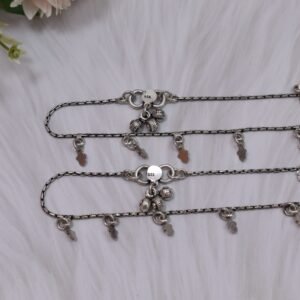 a pair of classic chained leaf shape drop 925 sterling silver anklets stretched across a white textured surface and white faux fur, featuring flat leaf charms and ghungroo bell clasps next to floral accents.