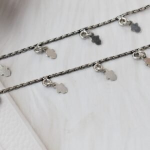 a pair of classic chained leaf shape drop 925 sterling silver anklets stretched across a white textured surface and white faux fur, featuring flat leaf charms and ghungroo bell clasps next to floral accents.