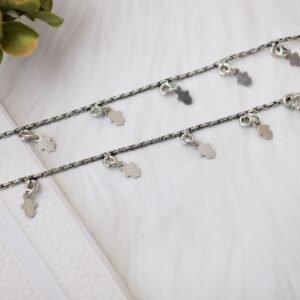 a pair of classic chained leaf shape drop 925 sterling silver anklets stretched across a white textured surface and white faux fur, featuring flat leaf charms and ghungroo bell clasps next to floral accents.