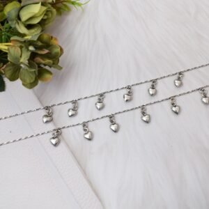 Full-length view of two chained heart drop silver anklets stretched out on a white surface next to floral accents, displaying the entire chain length, evenly spaced hearts, and end clasps with small bells.