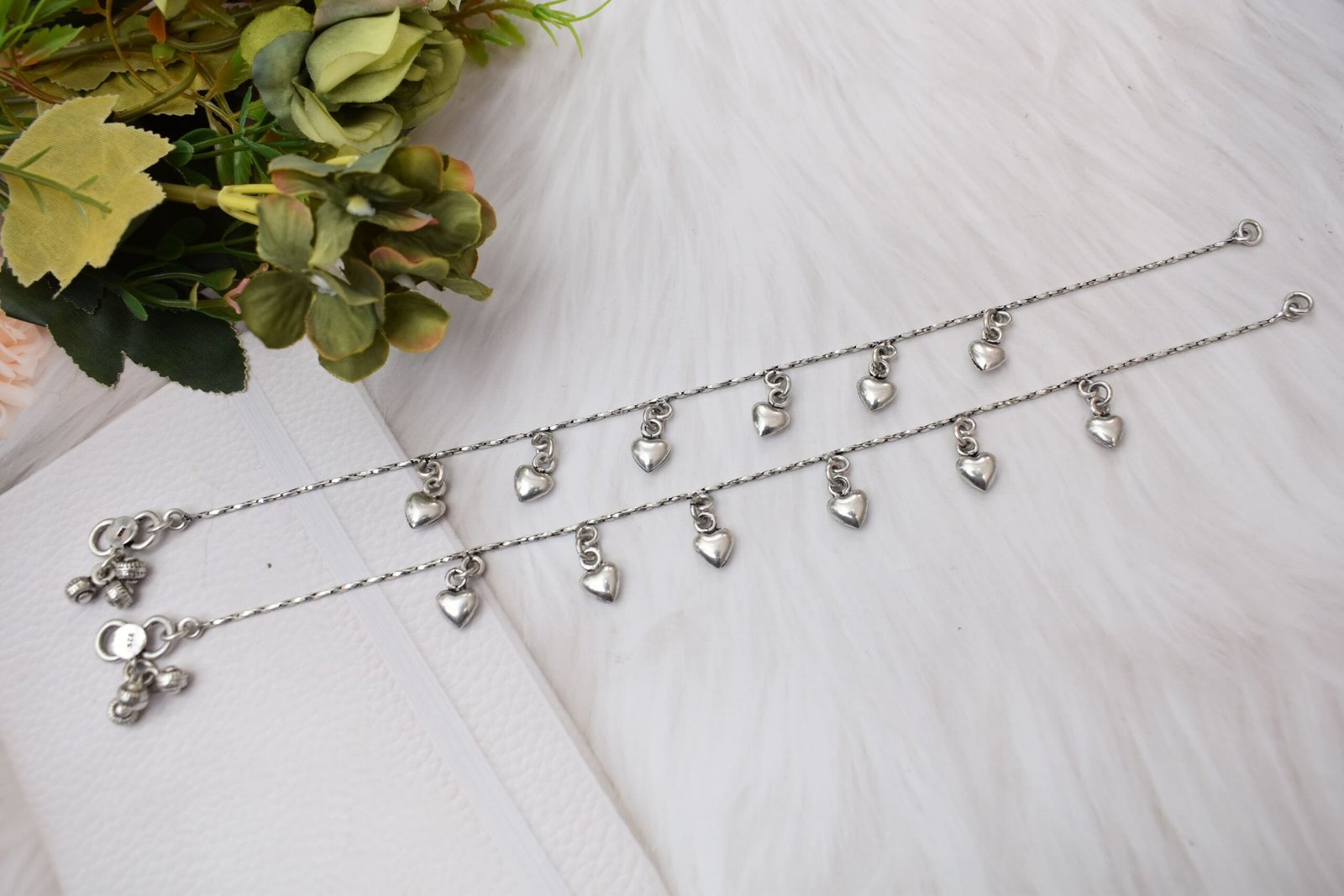 desireable-chained-heart-drop-silver-anklets1 Full-length view of two chained heart drop silver anklets stretched out on a white surface next to floral accents, displaying the entire chain length, evenly spaced hearts, and end clasps with small bells.